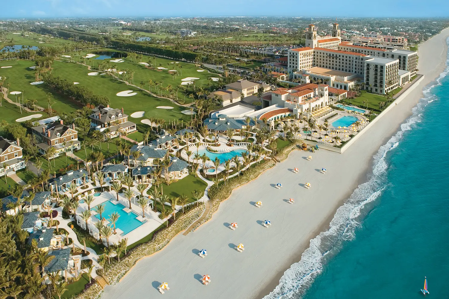 Ein Golfplatz und eine große Hotelanlage mit rötlichen Dächern, mehreren Pools und Palmen liegen an einem weißen Sandstrand, dahinter das türkisfarbene Meer