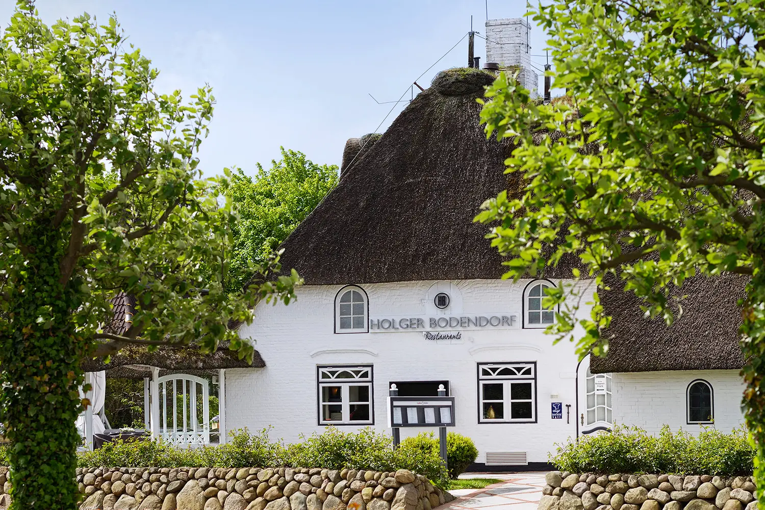 Ein weiß getünchtes Haus mit reetgedecktem Dach und dunklen Sprossenfenstern, davor grüne Bäume und eine niedrige Natursteinmauer, blauer Himmel