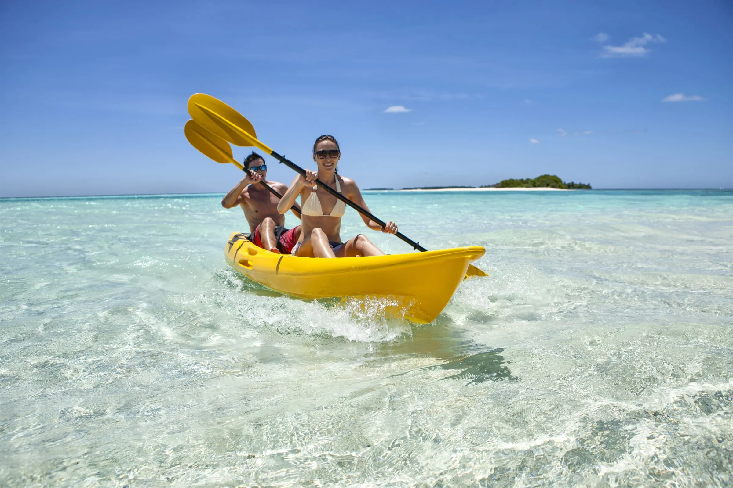 Ein Mann und eine Frau paddeln in einem gelben Kajak durch klares, flaches Wasser, im Hintergrund eine kleine Insel mit Strand und Bäumen unter blauem Himmel