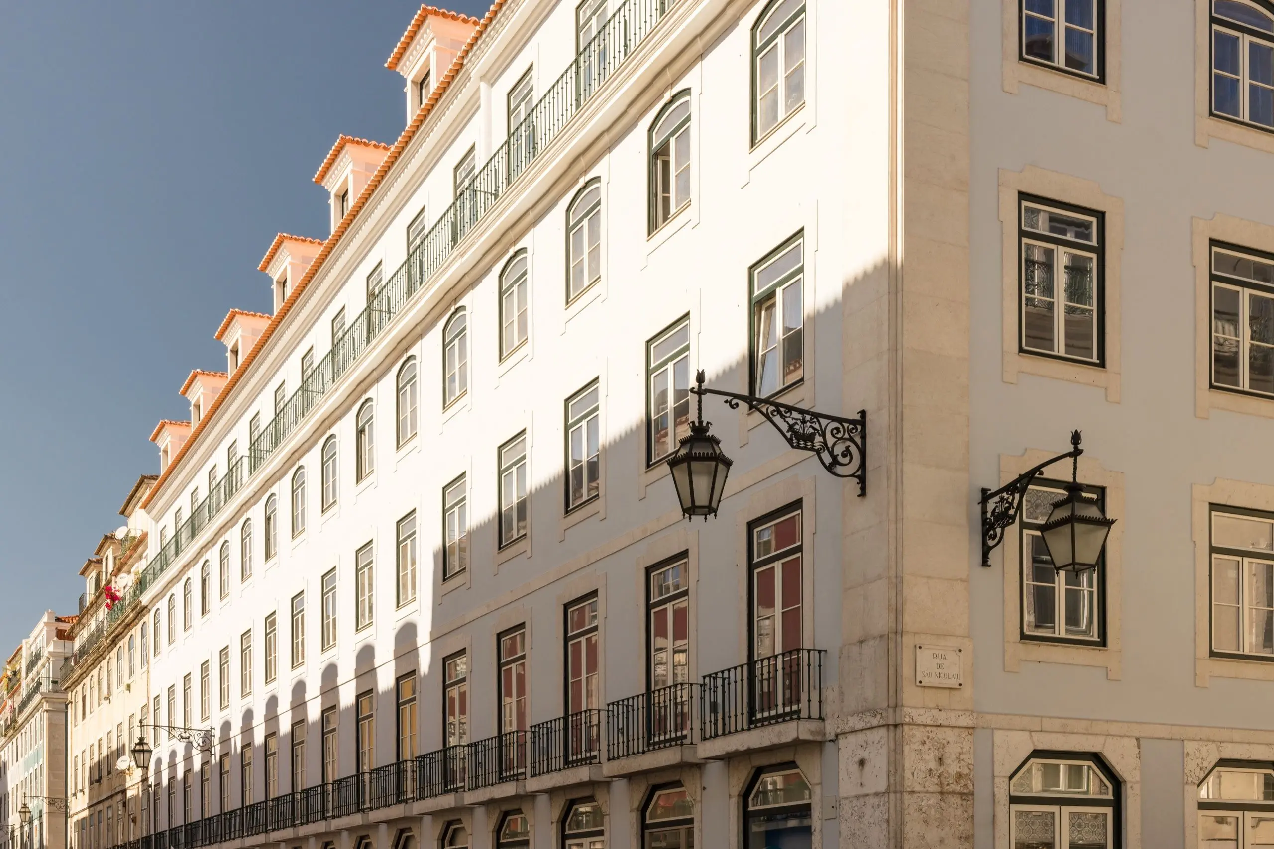 Eine helle Hausfassade mit Dachgauben, Balkonen und orangefarbenen Dachziegeln unter blauem Himmel, an der Ecke zwei schmiedeeiserne Straßenlaternen im Sonnenlicht