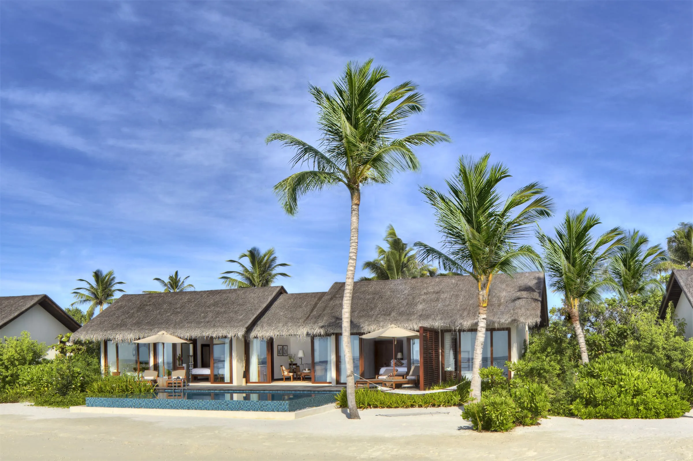 Eine Villa mit Palmdach und offenen Glastüren, davor ein Pool, Sonnenschirme und heller Sand, ringsum hohe Palmen und Büsche unter blauem Himmel
