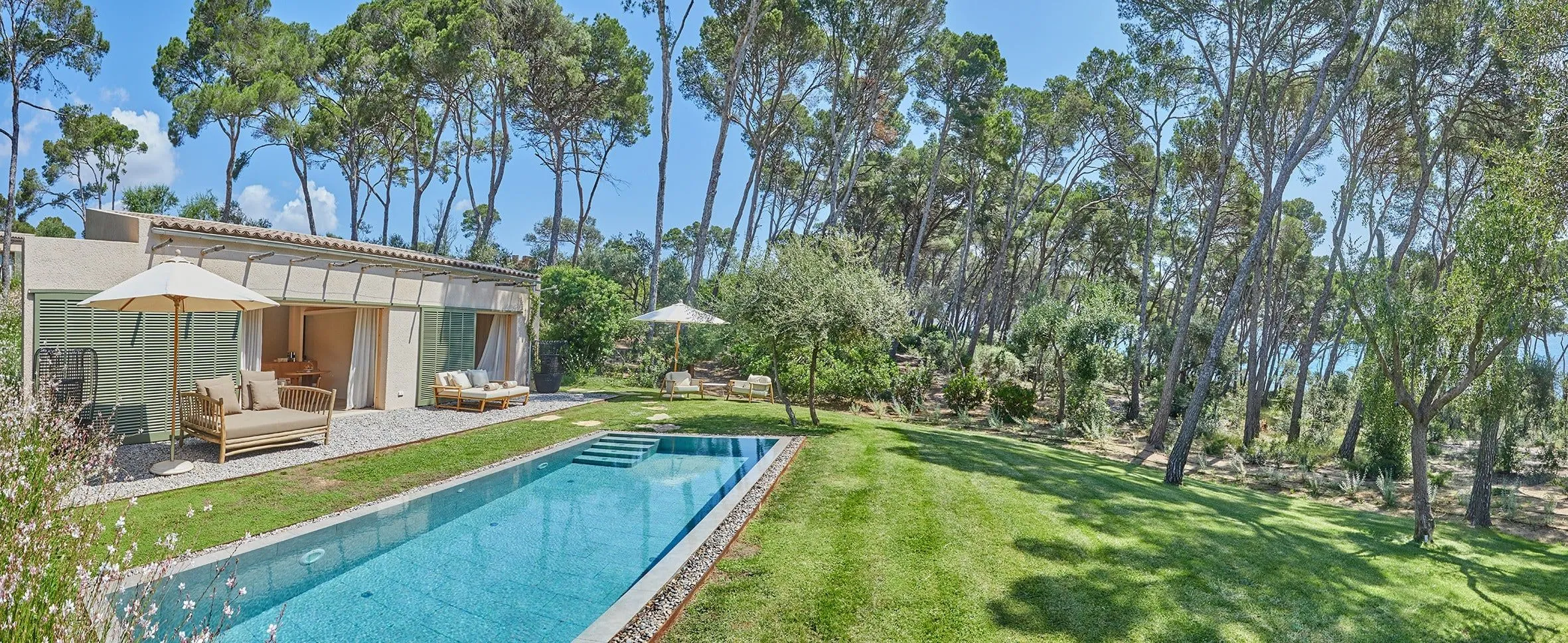 Langer Pool vor einem flachen Haus mit Terrasse, Sofas und Sonnenschirmen, daneben Rasen und hohe Pinien, zwischen den Stämmen das Meer