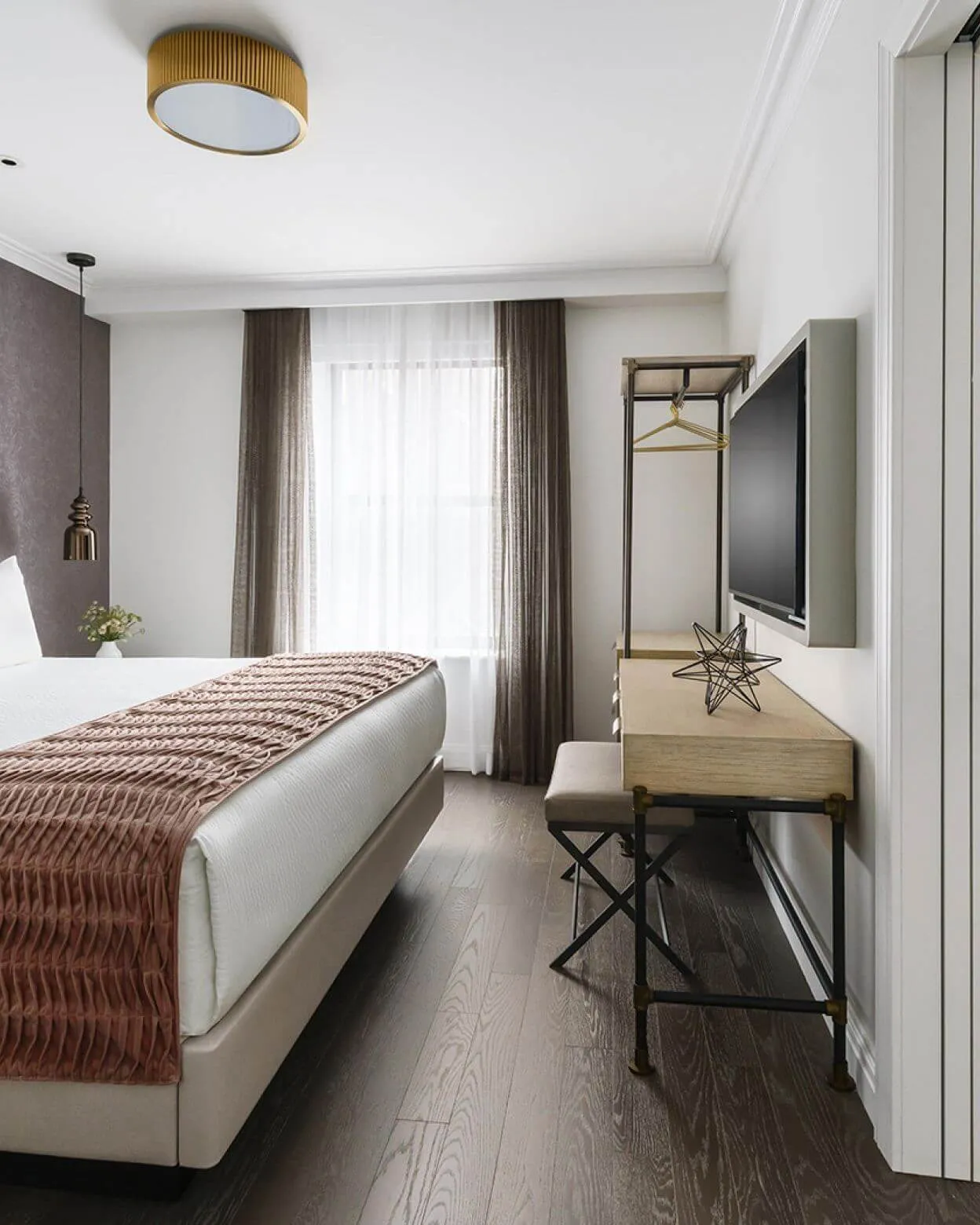 Schlafzimmer mit Bett und rostrotem, plissiertem Überwurf, messingfarbener Deckenleuchte und Pendelleuchte, rechts ein Holzschreibtisch mit Hocker, ein offener Kleiderständer mit Bügel und ein Wandfernseher, hinten ein Fenster mit Vorhängen