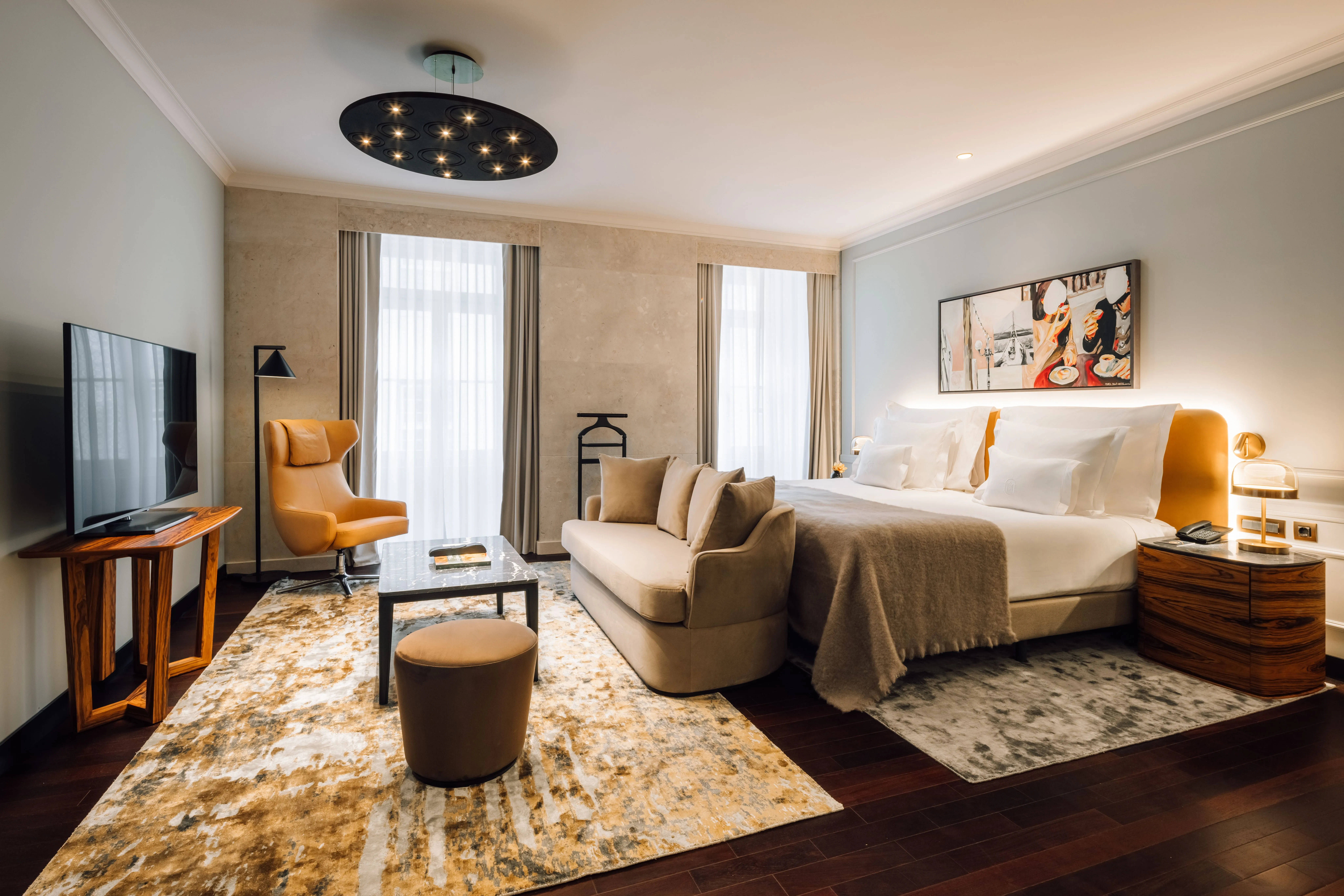 Ein Schlafzimmer mit breitem Bett, Kissen und Überwurf, darüber ein Bild, davor ein Sofa, links ein Sessel, Fernseher und Couchtisch auf gemustertem Teppich, an der Decke eine runde dunkle Leuchte mit vielen Lichtpunkten, hinten zwei Fenster mit Vorhängen
