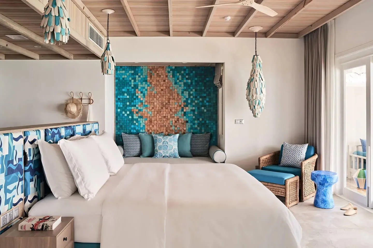 Schlafzimmer mit großem Bett und blau gemustertem Kopfteil, einer Sitznische vor einer Mosaikwand in Türkis und Kupfer, Hängeleuchten aus Holzplättchen und einem Korbsessel am Fenster