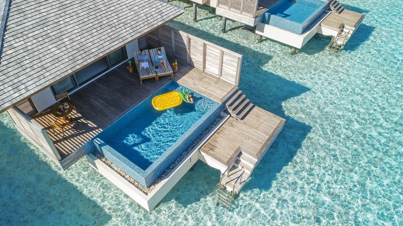 Eine Wasservilla mit Schindeldach von oben: auf dem Holzdeck liegen zwei Menschen auf Liegen, im Pool liegt ein Mensch auf einer grünen Luftmatratze neben einer gelben, Treppen und Leitern führen ins klare, türkisfarbene Wasser, oben rechts der Pool der Nachbarvilla