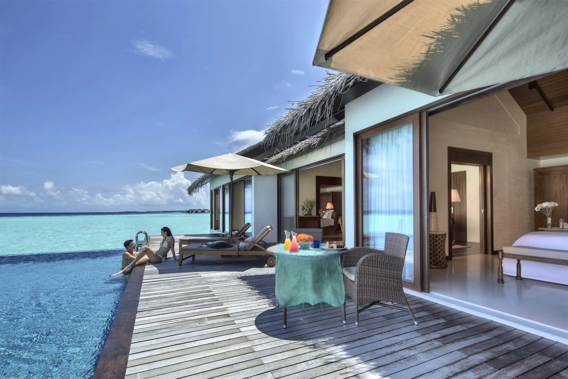 Holzdeck einer Villa über der Lagune mit Pool, Liegen unter einem Sonnenschirm und einem Tisch mit Karaffen, orangefarbenen Blumen und einem Obstteller, am Poolrand sitzen ein Mann und eine Frau, rechts der Blick durch offene Glastüren auf ein Bett