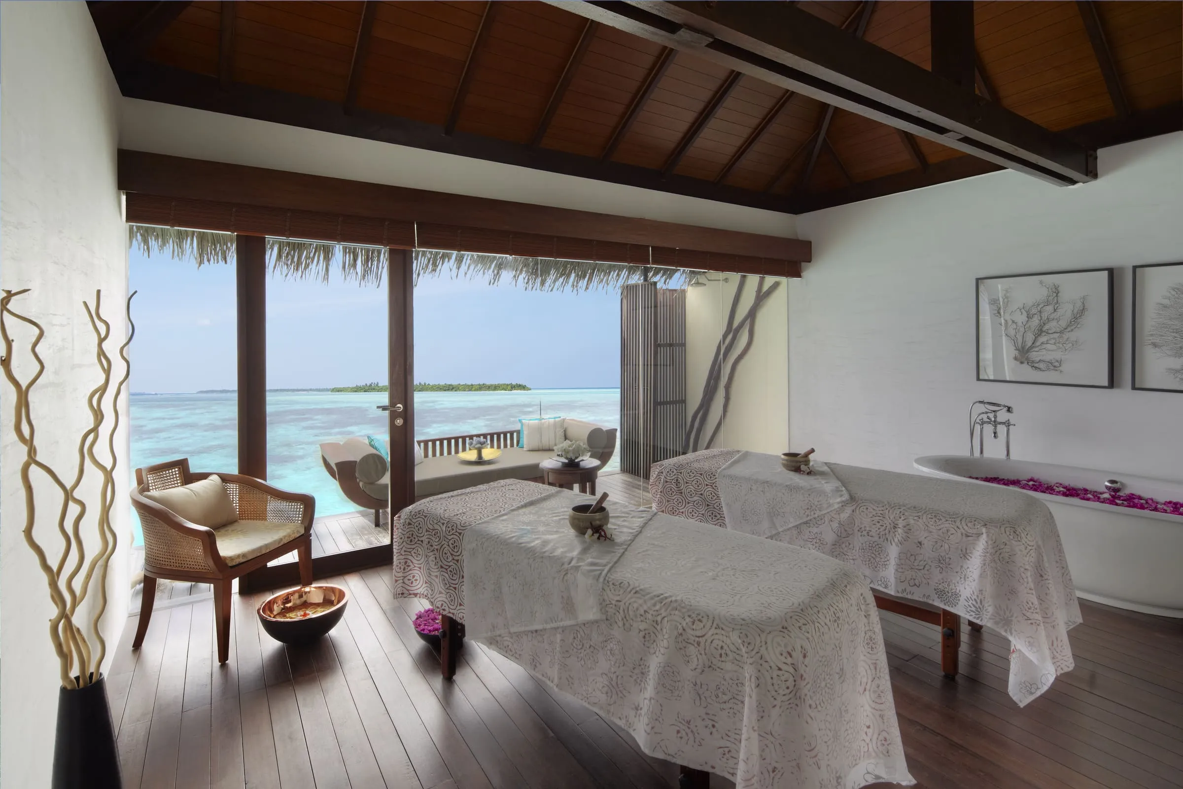 Behandlungsraum mit zwei Massageliegen unter Spitzendecken, rechts eine freistehende Wanne mit Blütenblättern, links ein Rattansessel und eine offene Glasfront zu einer Terrasse mit Sofa über der Lagune, am Horizont eine bewaldete Insel