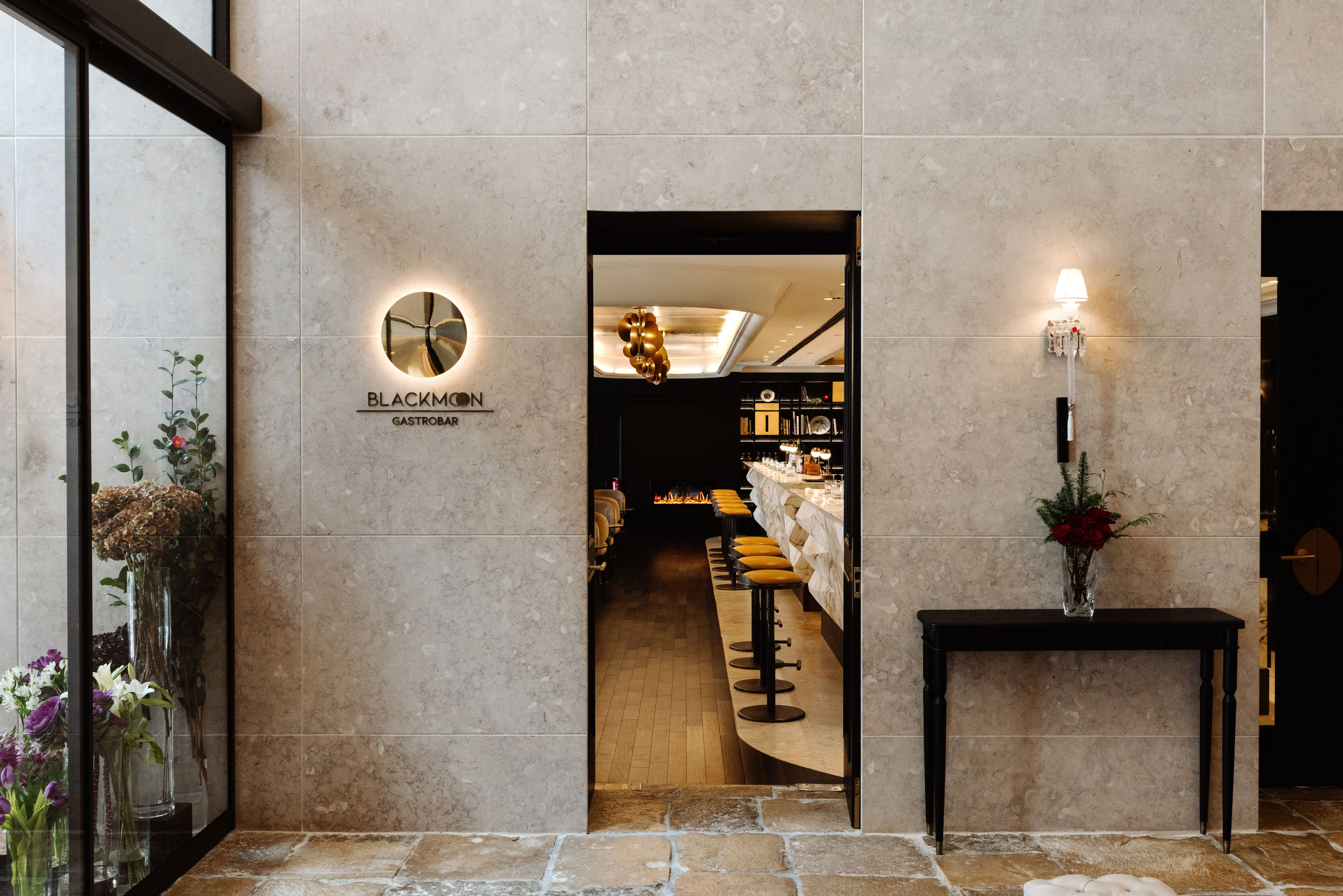 Eine Wand aus hellen Steinplatten mit dem beleuchteten Schild BLACKMOON Gastrobar neben einem offenen Durchgang, dahinter eine Bar mit Marmortheke und Hockern, rechts ein schwarzer Konsolentisch mit roten Blumen, links eine Glaswand mit Blumen in Vasen