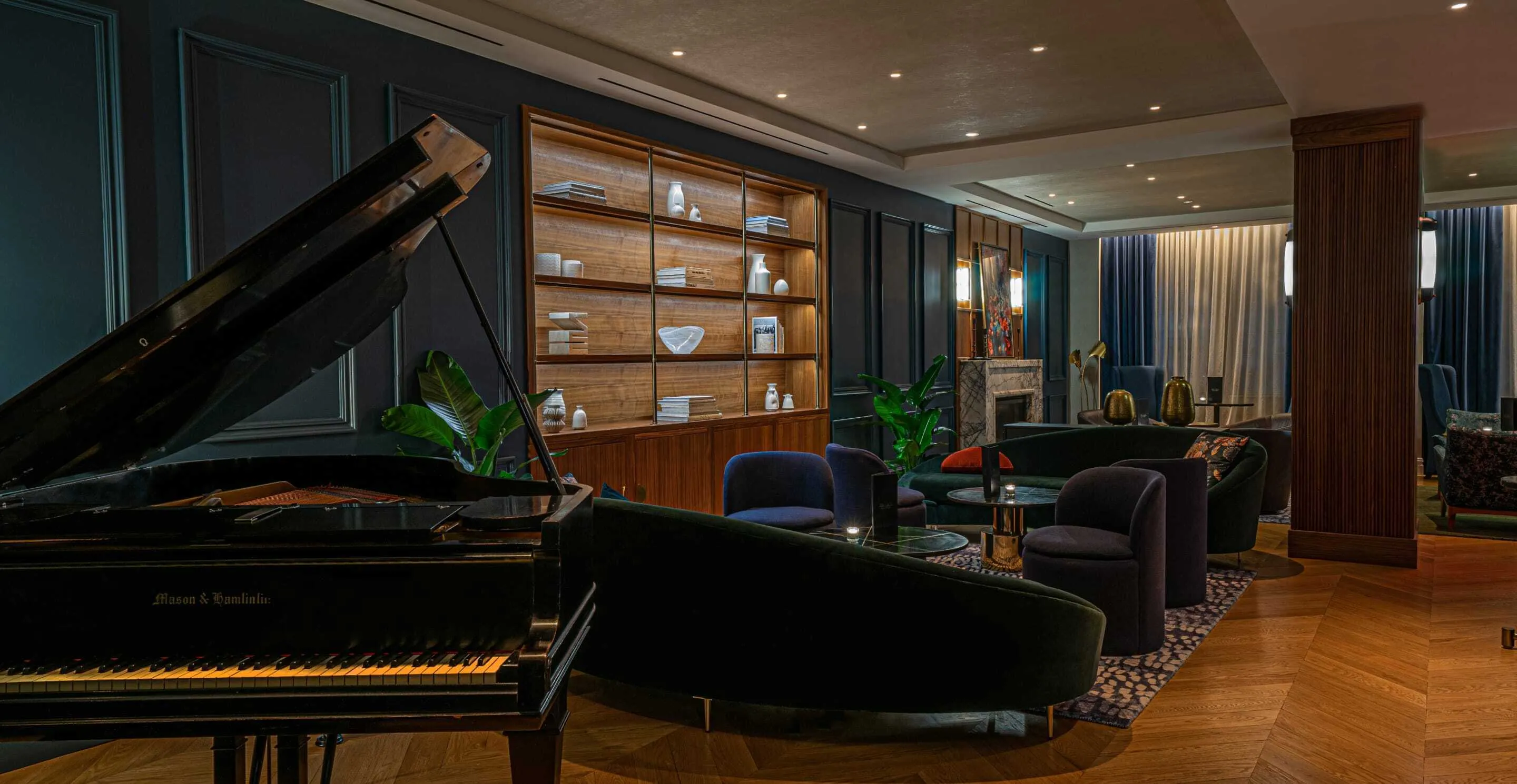 Gedämpft beleuchtete Lounge mit einem schwarzen, geöffneten Flügel im Vordergrund, dahinter ein geschwungenes Sofa, Samtsessel, niedrige Tische, ein beleuchtetes Holzregal und ein Kamin, an den Fenstern blaue Vorhänge