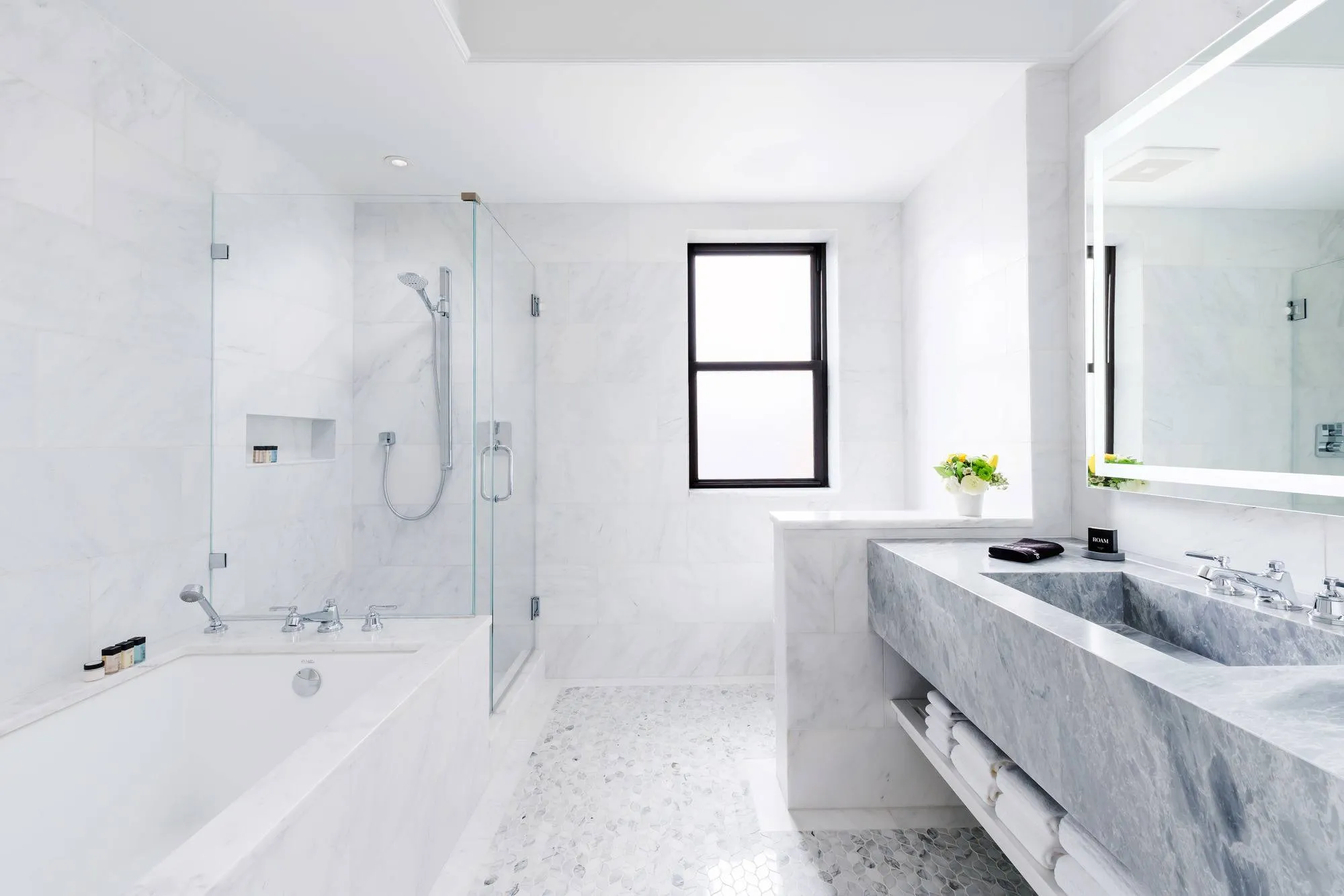 Bad aus weißem Marmor mit Badewanne, Glasdusche und einem Fenster, rechts ein Waschtisch aus grauem Marmor mit Spiegel und gestapelten Handtüchern darunter, auf dem Boden kleine Mosaikfliesen