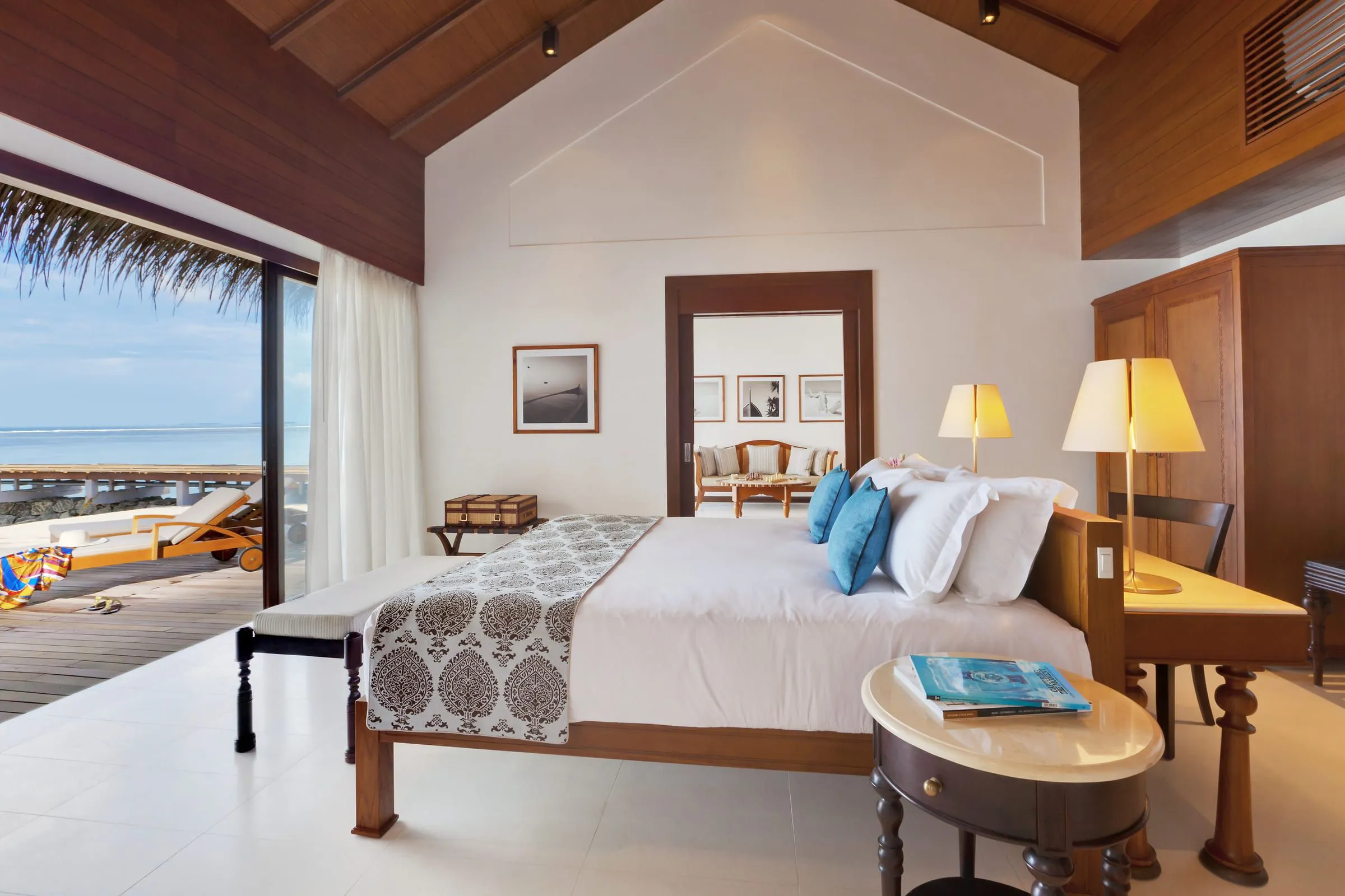 Schlafzimmer unter einer holzverkleideten Dachschräge mit großem Bett, blauen Kissen und gemustertem Überwurf, links eine offene Glastür zum Holzdeck mit Liege und Meer, hinten eine Tür zum Wohnraum