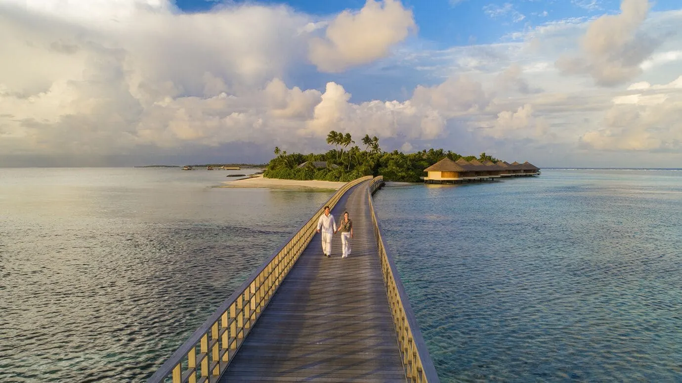 Zwei Menschen in heller Kleidung gehen über eine lange Holzbrücke auf eine Insel mit Palmen, Sandstrand und einer Reihe runder Wasservillen zu, darüber große Wolken