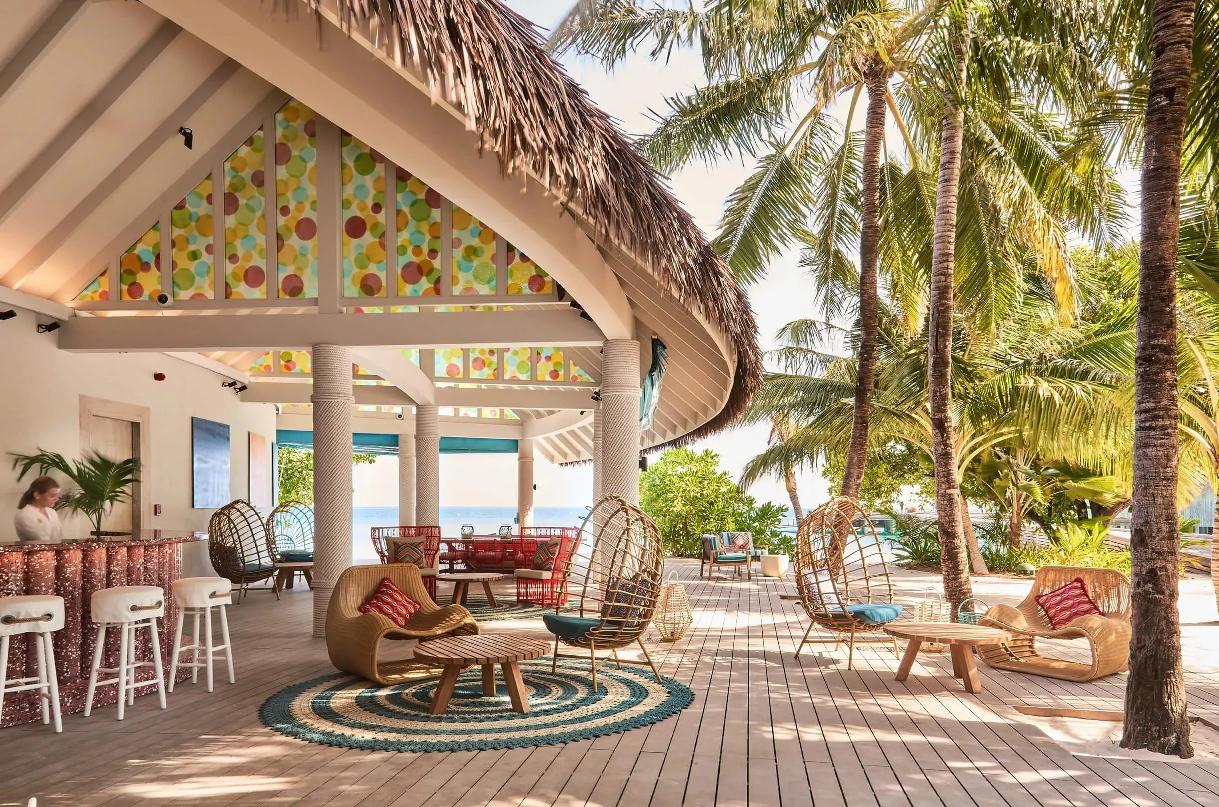 Offener Pavillon mit Reetdach und bunten Glasfenstern, darunter eine Bar mit Hockern, Korbsessel auf einem Holzdeck, rechts Palmen und dahinter das Meer