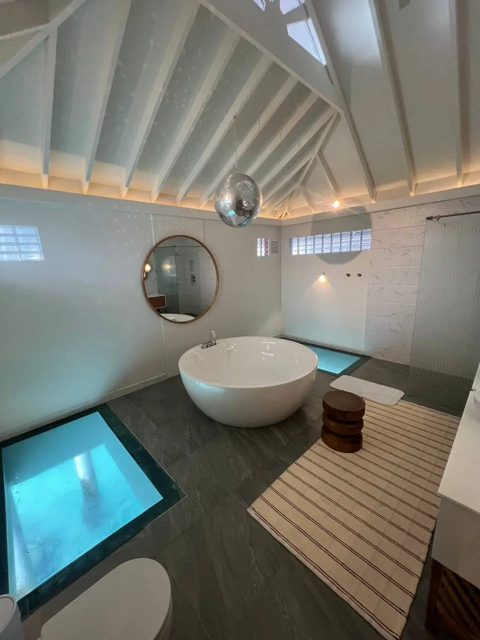 Über einer runden weißen Badewanne hängt eine Spiegelkugel, durch Fenster im Boden ist türkisfarbenes Wasser zu sehen.