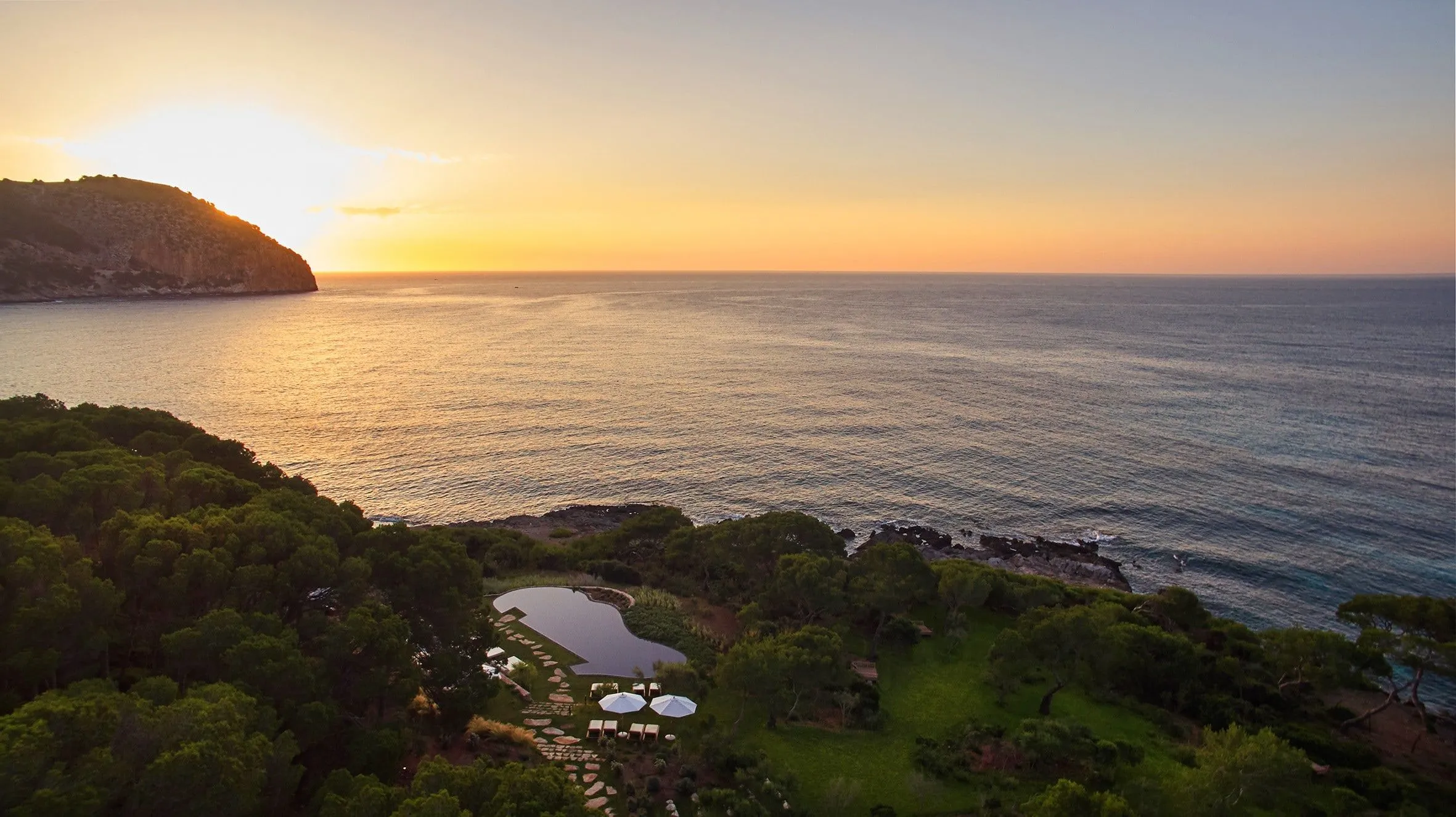 Küste mit Pinienwald, einem Pool mit weißen Schirmen und Rasen, davor das Meer mit goldenem Glanz, links eine Landzunge und die tief stehende Sonne