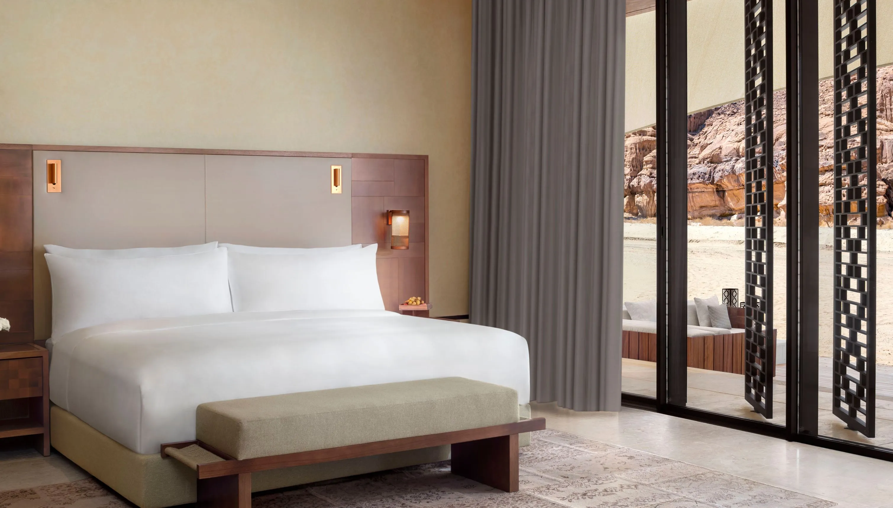Ein Schlafzimmer mit breitem, weiß bezogenem Bett, gepolsterter Bank am Fußende und grauem Vorhang, rechts raumhohe Glastüren mit dunklen Gitterpaneelen, draußen eine Terrasse, Sand und Sandsteinfelsen