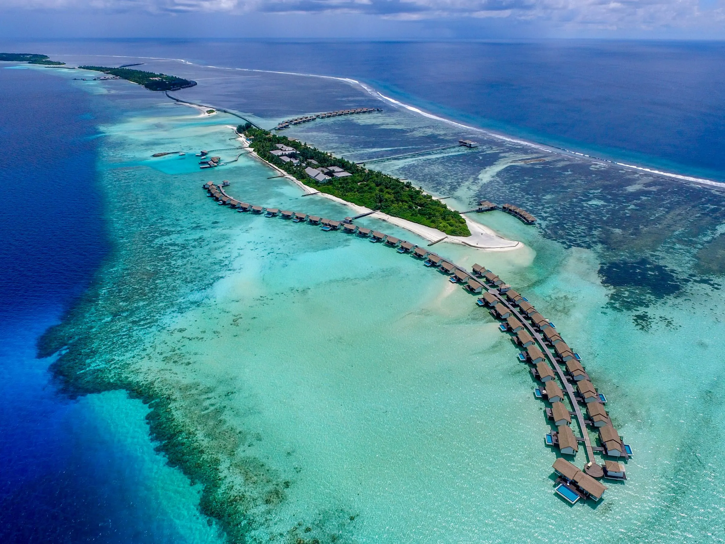 Lange Reihen von Wasserbungalows führen von einer bewaldeten Insel ins türkisfarbene Lagunenwasser, ringsum ein Riff mit Brandungslinie im tiefblauen Meer