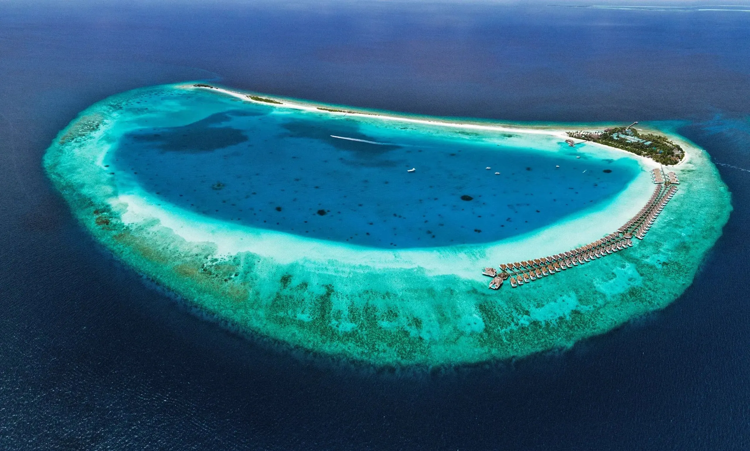 Ovales Atoll mit tiefblauer Lagune und türkisfarbenem Riffkranz im dunklen Ozean, an einem Ende eine bewachsene Insel und eine lange Reihe von Wasservillen