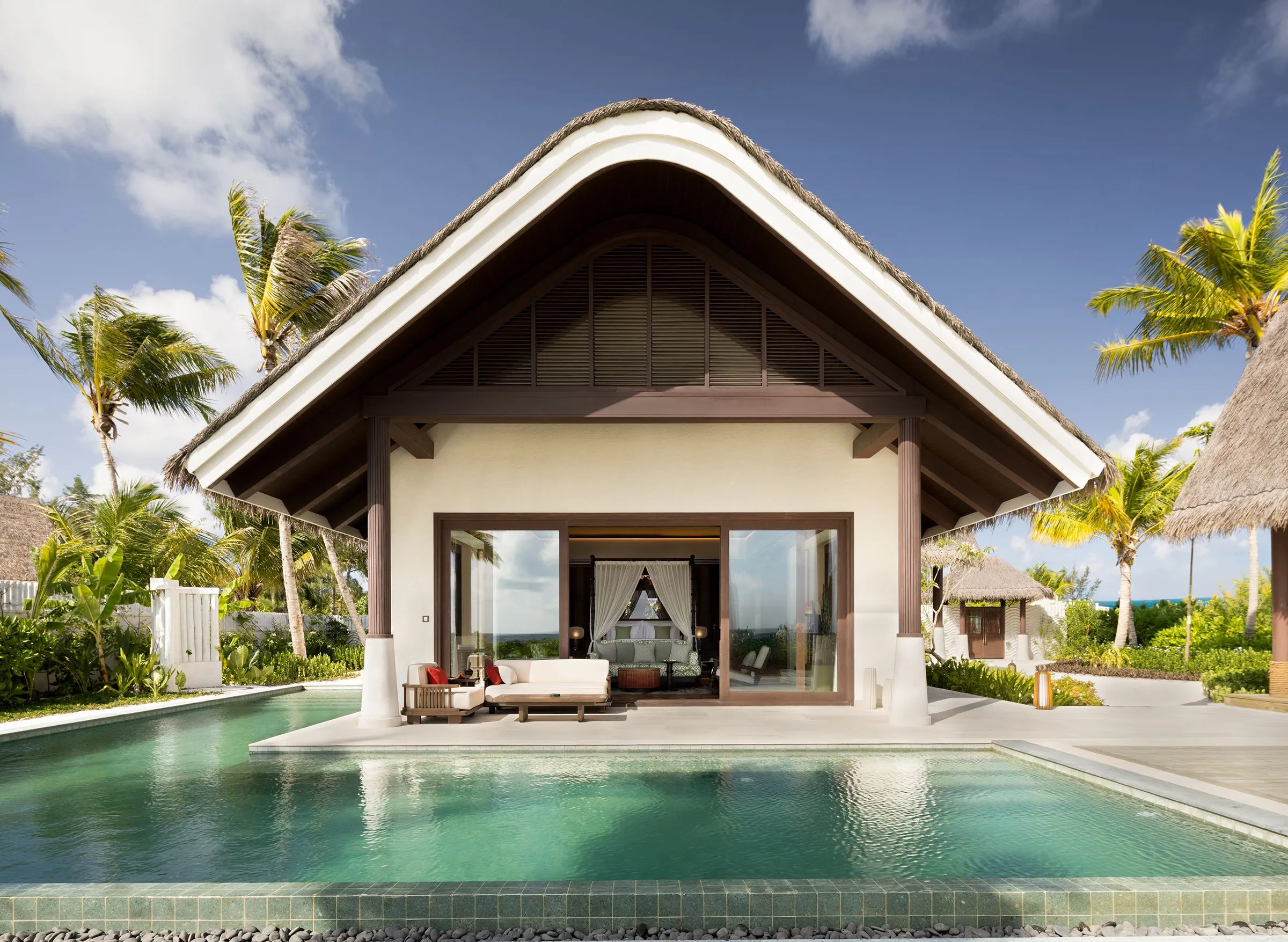 Ein Haus mit steilem Strohdach und Glasfront, davor eine Terrasse mit Sofa und Sessel und ein türkisfarbener Pool, ringsum Palmen und weitere strohgedeckte Häuser