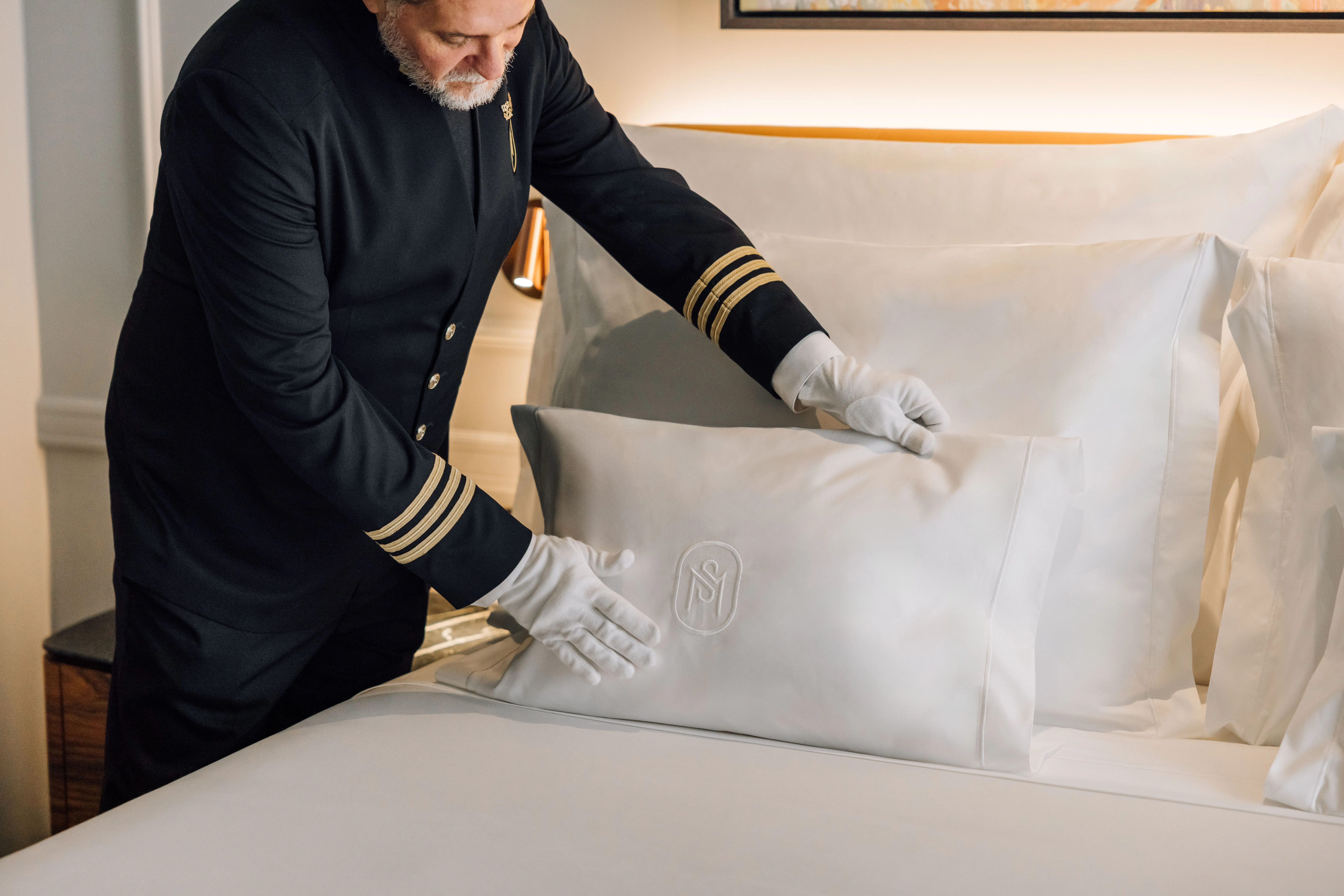 Ein Mann mit grauem Bart in dunkler Uniform mit goldenen Ärmelstreifen und weißen Handschuhen richtet ein Kissen mit gesticktem Monogramm auf einem weiß bezogenen Bett