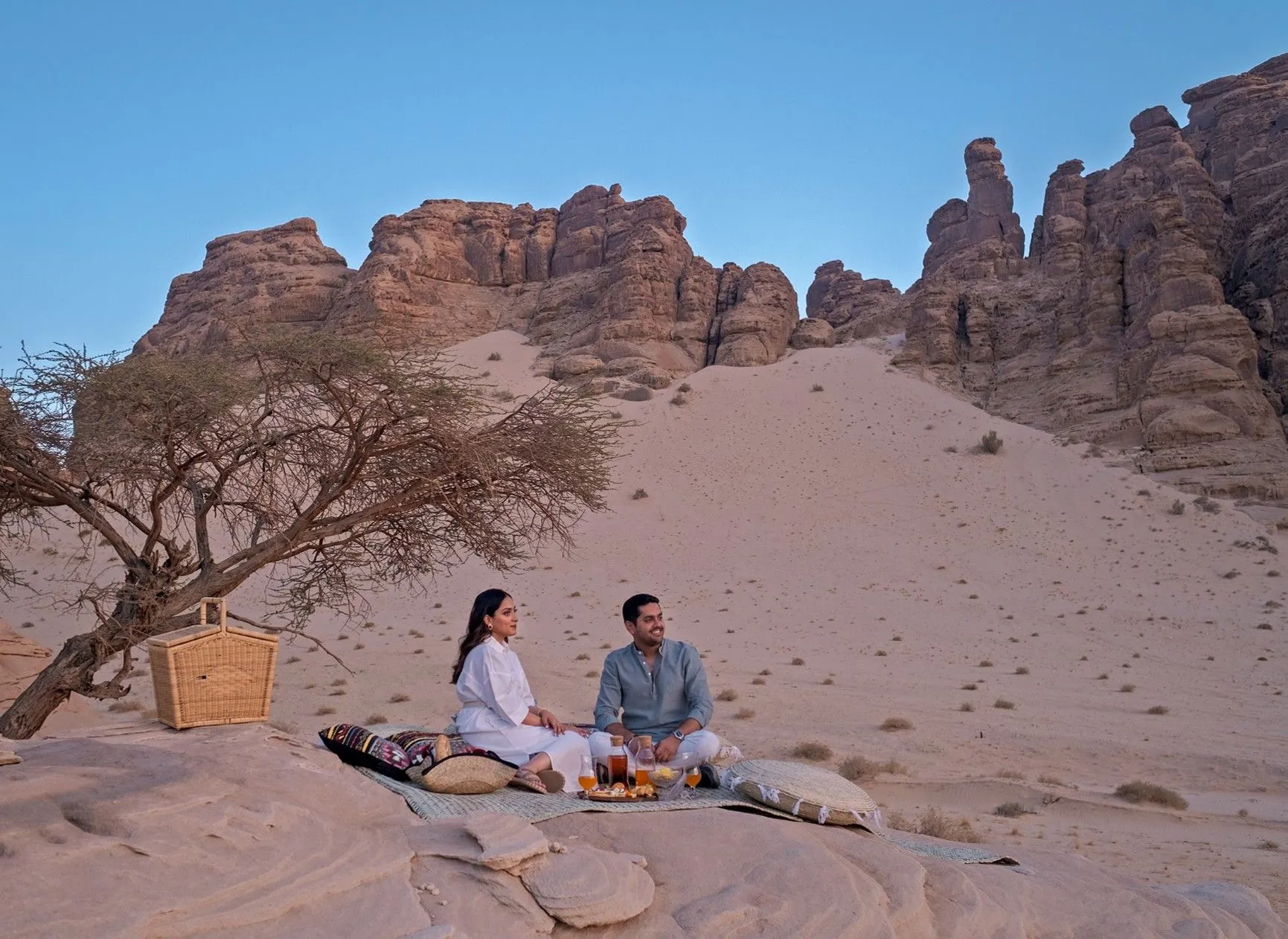 Eine Frau und ein Mann sitzen auf einer Decke auf flachem Fels, vor ihnen Getränke und Speisen, daneben Kissen, ein Picknickkorb und ein knorriger Baum, dahinter eine Sanddüne und zerklüftete Sandsteinfelsen unter blauem Himmel